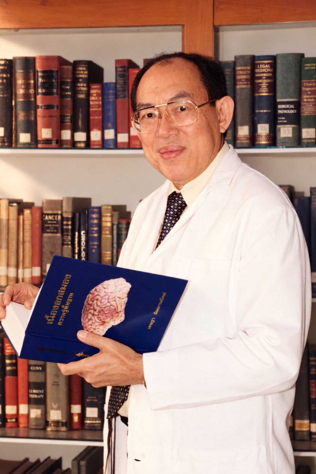 Associate Professor Dr. Jesada Nimmannitya in a white coat, holding his textbook in front of a bookshelf.